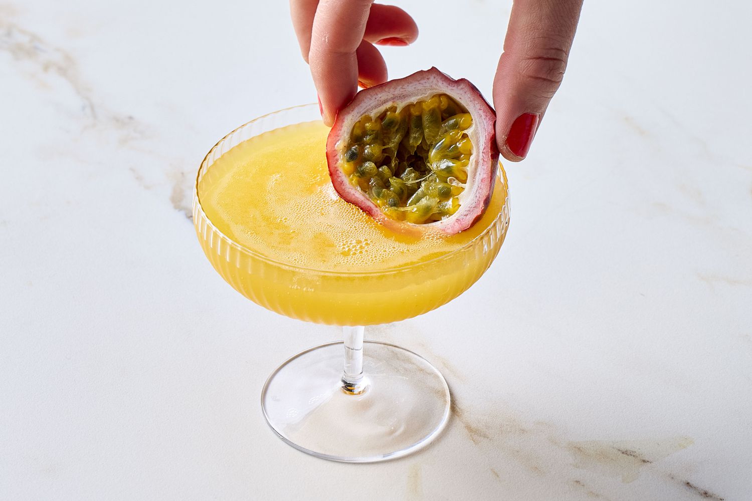 A passion fruit being placed onto the rim of a cocktail glass filled with a yellow beverage