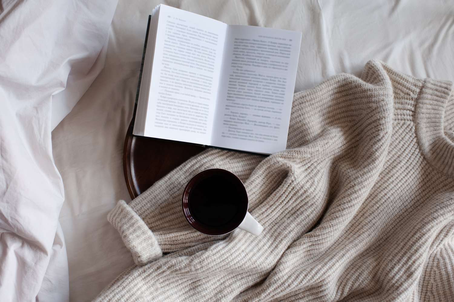Open paper book with cup of coffee on wooden tray and knit cloth textile in bed close up top view