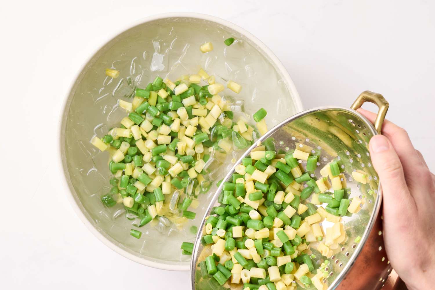 A hand pouring chopped green vegetables from a colander into a bowl of water