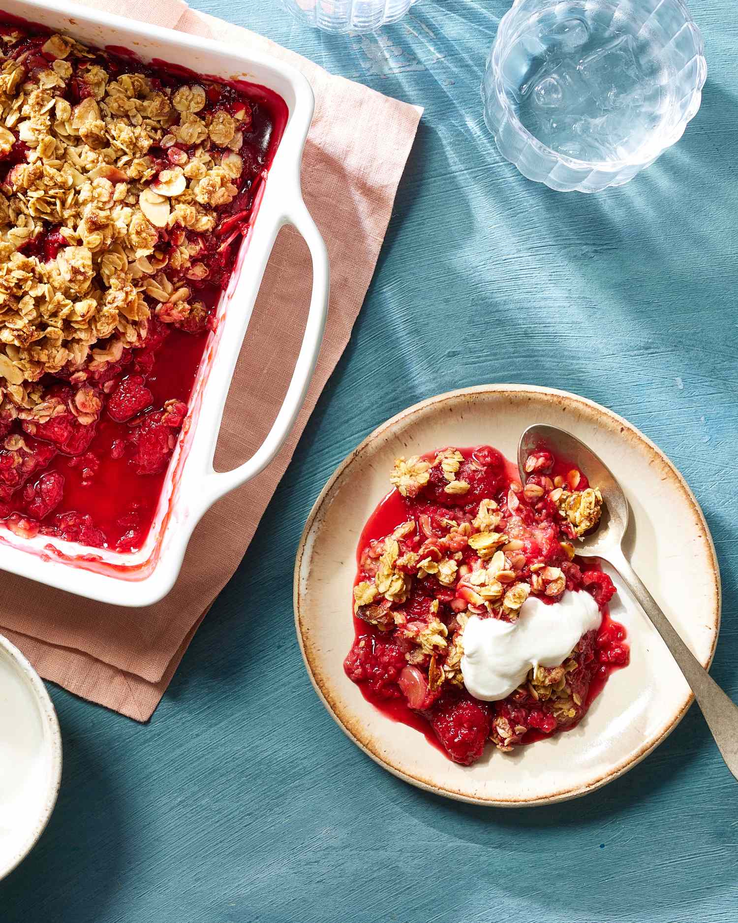 A raspberry almond crisp in a tray with a plated serving on a table, topped with cream and shown under natural light