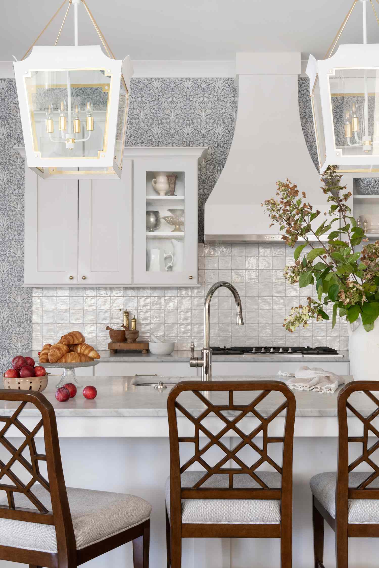 A kitchen with a white interior design, featuring a counter with wooden chairs, light fixtures, and decor items including fruit and flowers