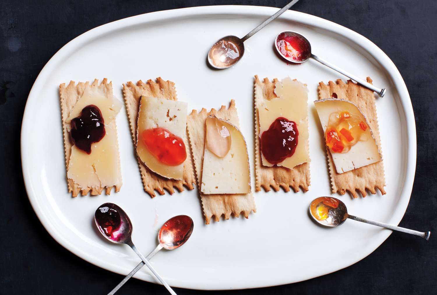 Crackers topped with slices of cheese and various fruit preserves arranged on a white plate with accompanying spoons of jam