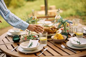 A wooden outdoor table set with dishes glasses floral decorations and food including corn and salad with a person reaching to arrange items