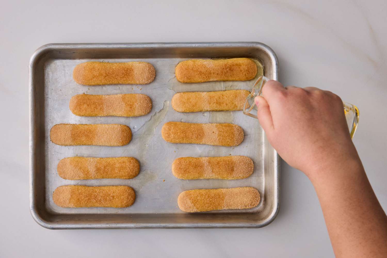 Hand pouring liquid over ladyfinger biscuits arranged on a baking sheet