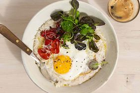 A bowl of savory oatmeal topped with a fried egg greens and cherry tomatoes accompanied by a cup of coffee