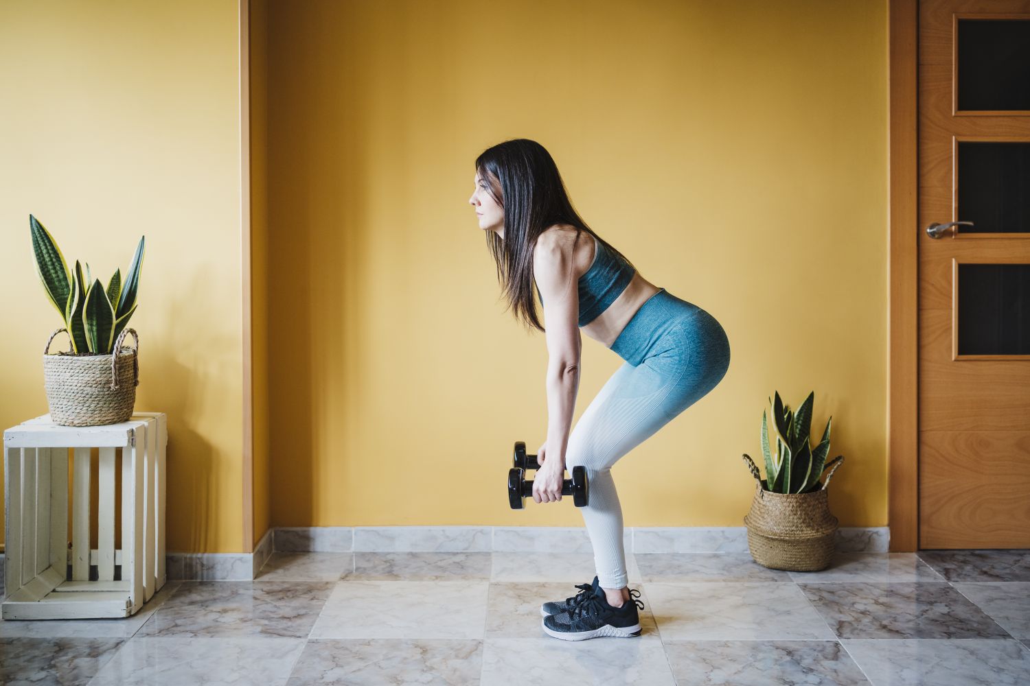 A person exercising at home with dumbbells performing a bentover movement in a room with decorative plants