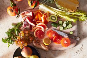 A variety of cut fruits and vegetables on a wooden board, including apples, citrus, celery, pineapple, carrots, grapes, and cucumber
