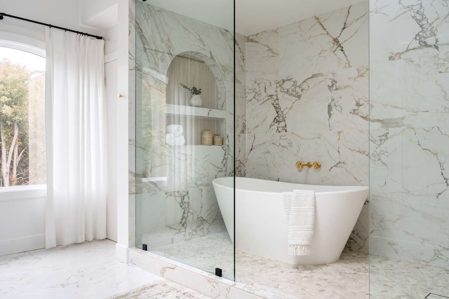 A modern bathroom with a freestanding bathtub, marble walls and flooring, a built-in shelf, and glass partition