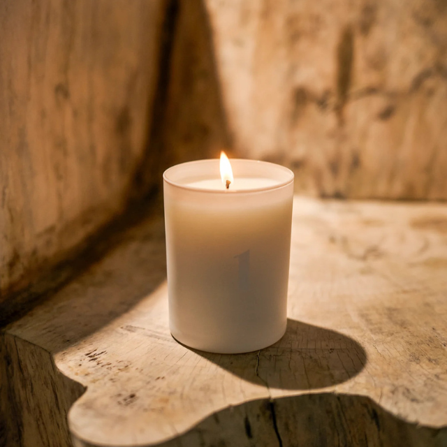 A lit candle in a simple holder set on a wooden surface casting a shadow