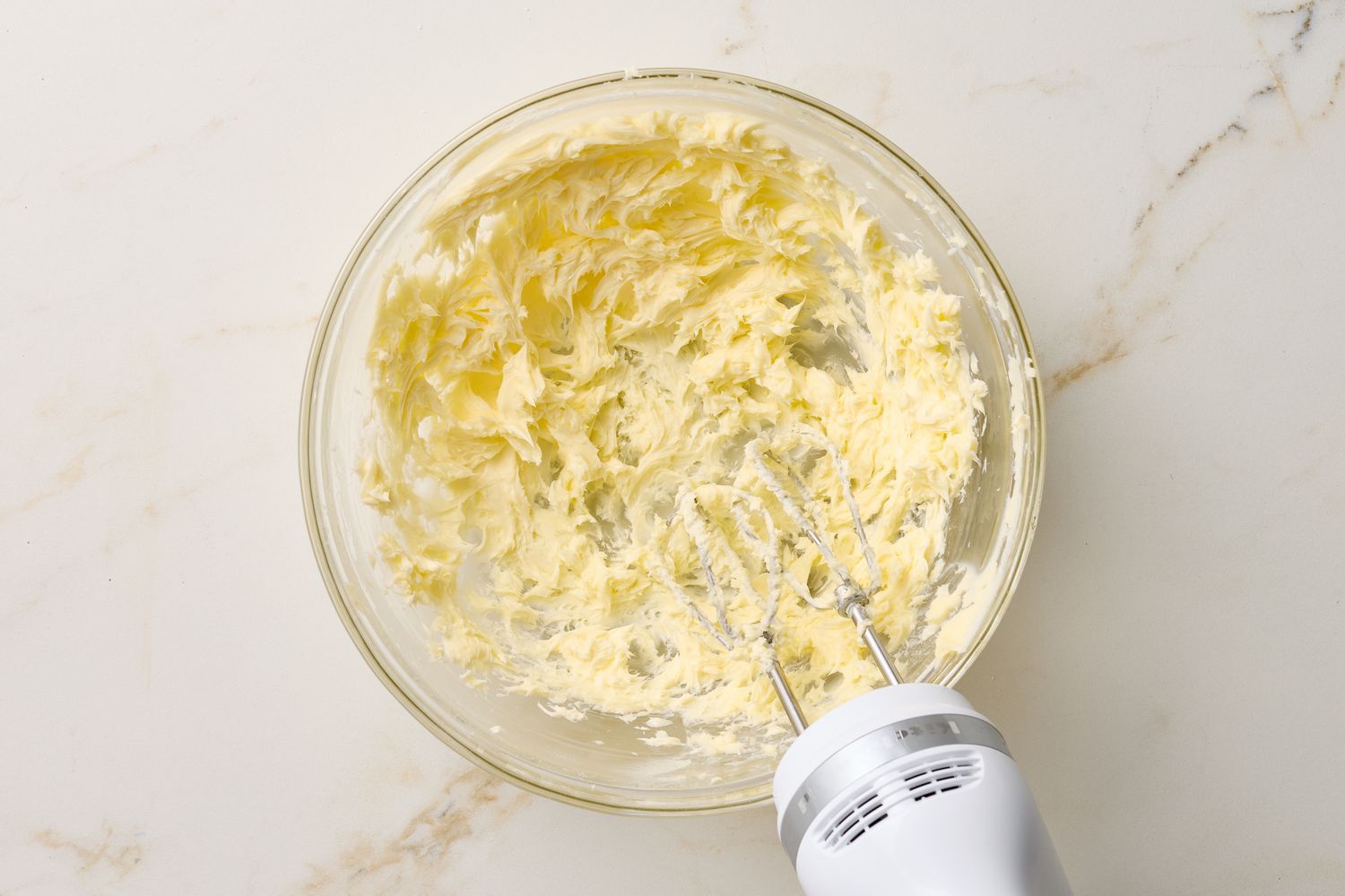 A glass bowl with creamed butter alongside a stand mixer with beaters inserted