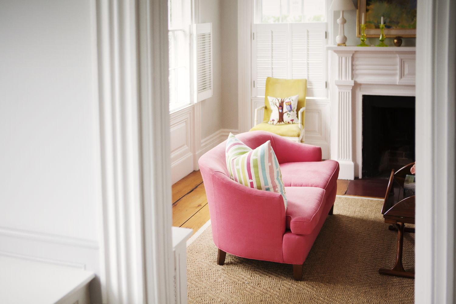 A cozy living room with a pink sofa a yellow chair and a fireplace featuring natural light through windows with shutters