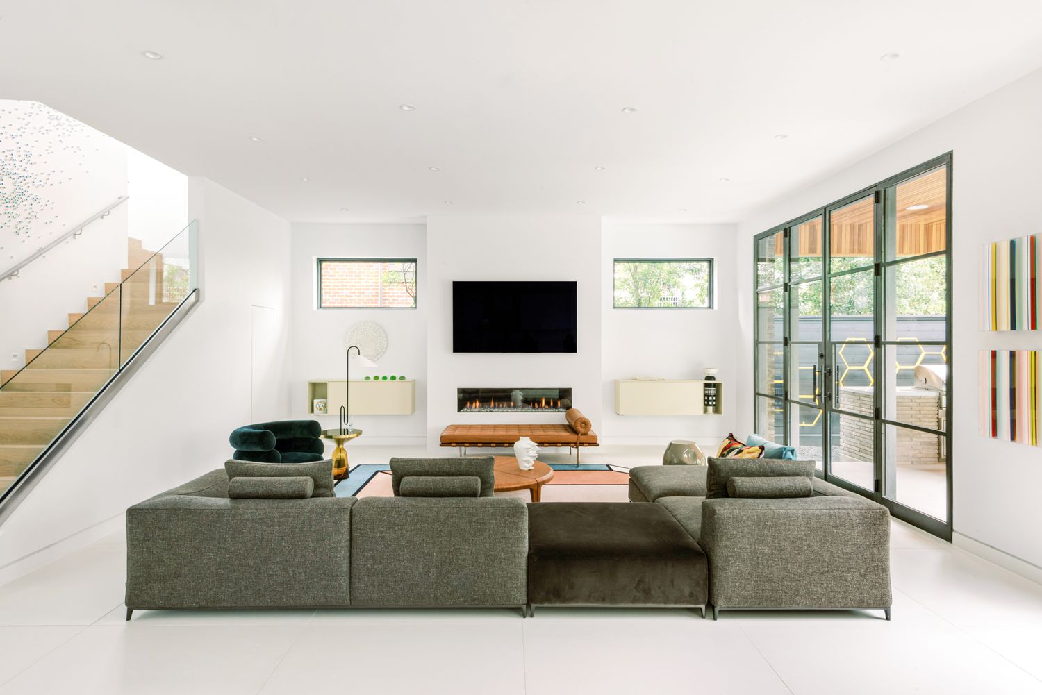 Minimalist living room by Lambrakos Studio: gray sectional sofa with velvet cushions, wood-and-glass coffee table, linear fireplace, and abstract art. Large windows with glass doors, white walls, light wood stairs, and natural light create a calm, stylish space.