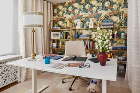 A desk with various items in a neatly arranged office space with bookshelves and decorative wallpaper in the background