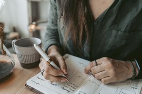 A person writing in a notebook with a pen a mug and desk items in view