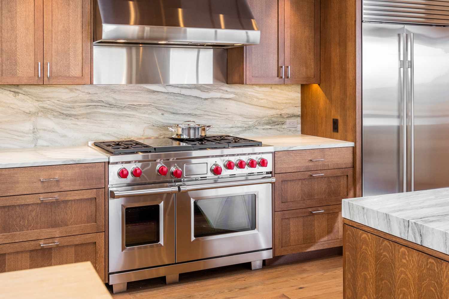 A modern kitchen featuring a stainless steel double oven range with red knobs and a vent hood installed within wood cabinetry