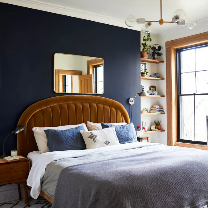A styled bedroom featuring a bed with a curved upholstered headboard pillows and blanket a shelf with decor items and a window