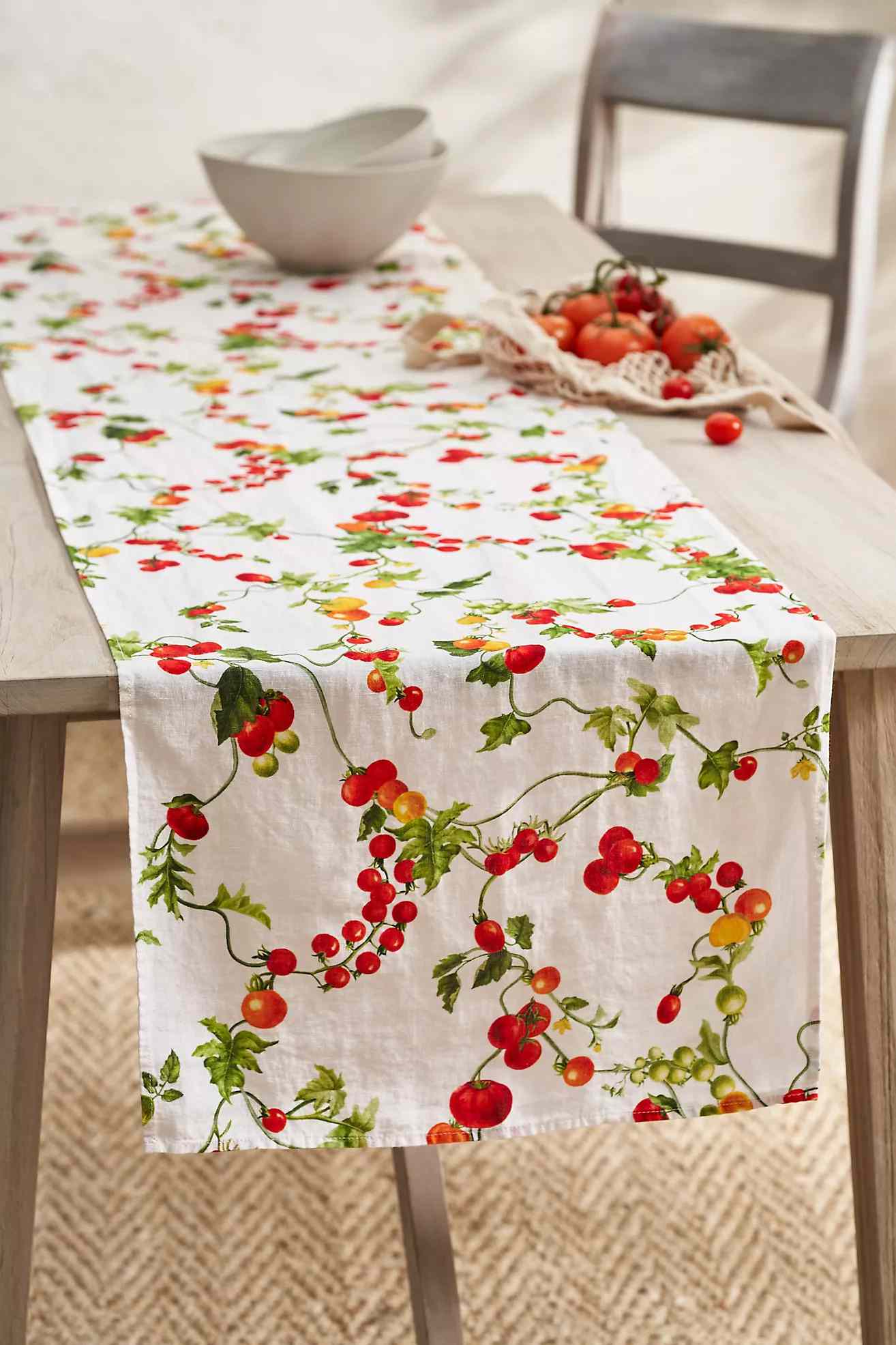 A decorated table runner with a tomato print on a dining table, accompanied by a wooden chair, a white bowl, and a basket of tomatoes