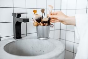 A hand reaching for a container of makeup brushes placed on a bathroom counter