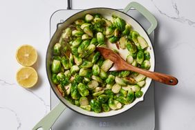 Brussels sprouts being sauted in a skillet with a wooden spatula nearby halved lemons placed on the side