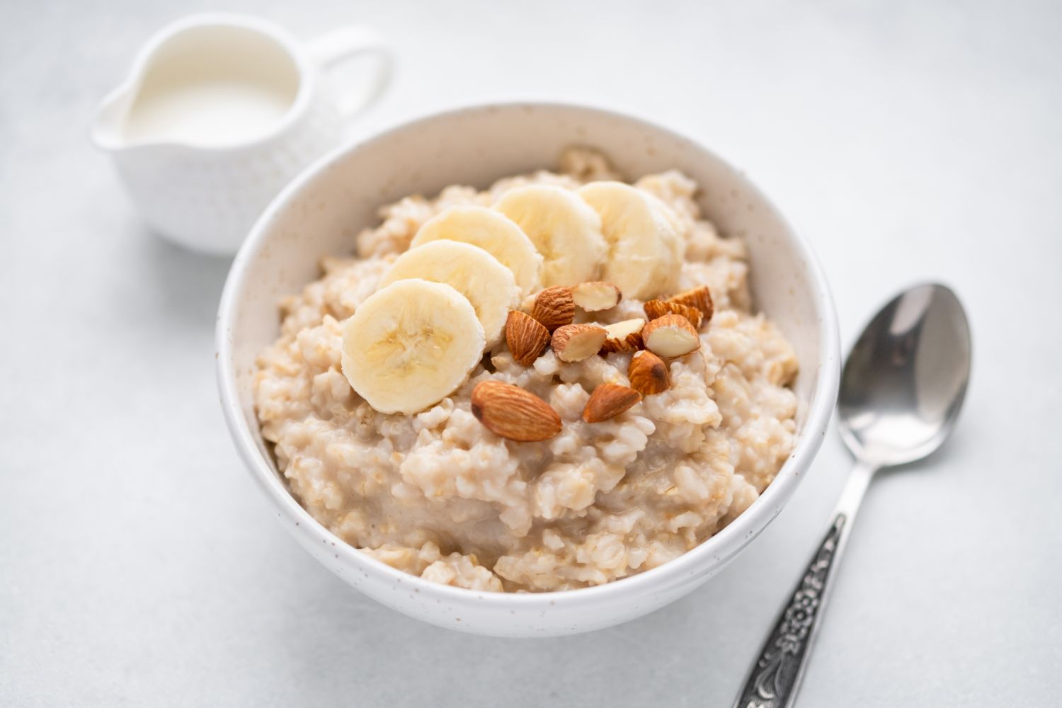 A bowl of oatmeal topped with banana slices and nuts spoon nearby and small pitcher in background