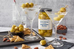 Two martini glasses garnished with olives a jar of blue cheese stuffed olives crackers almonds and a drink in the background