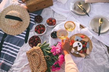 Picnic setup with food and drinks on a blanket outdoors featuring two glasses of a beverage being held up in a toast