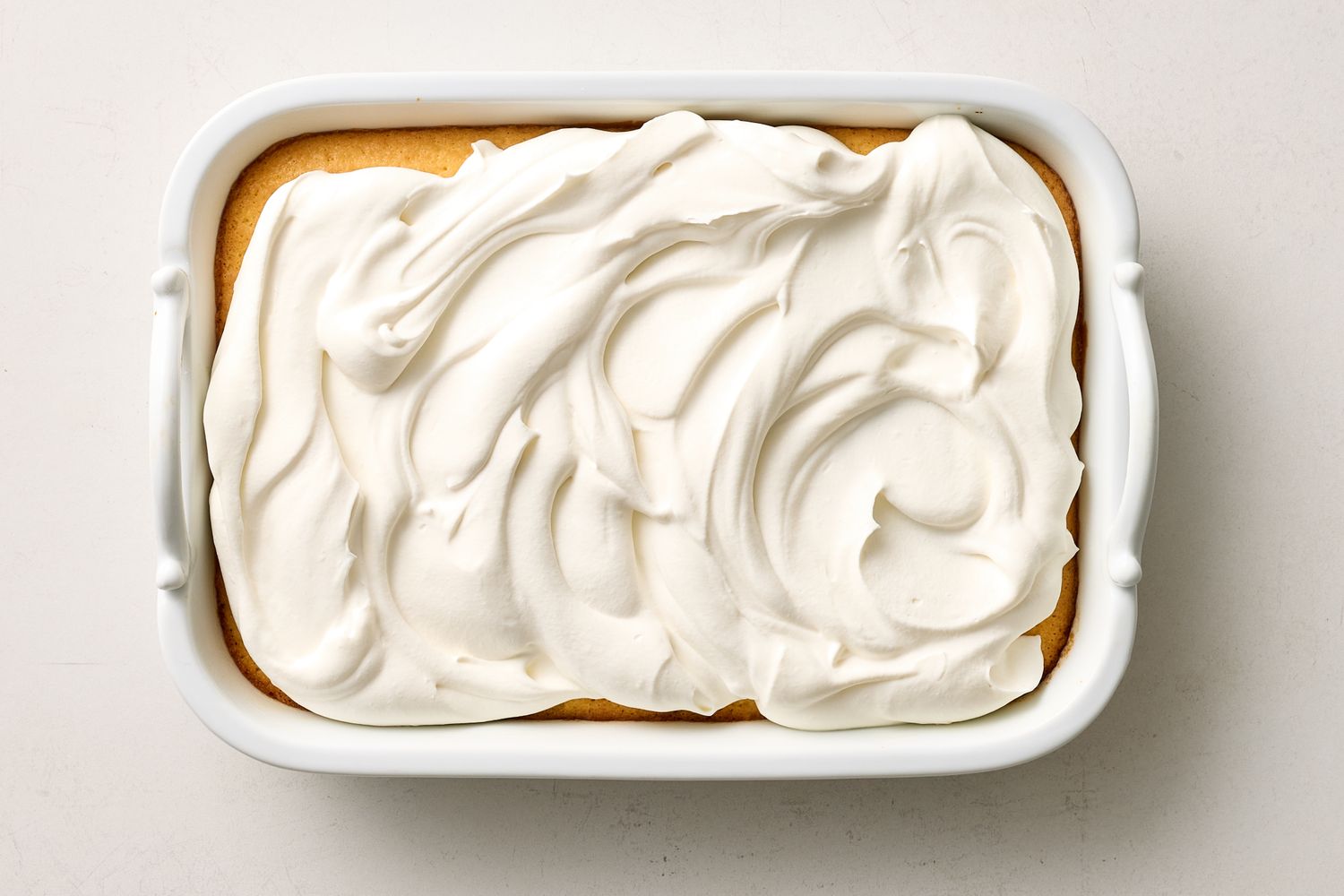 A rectangular dish with tres leches cake topped with whipped cream spread evenly