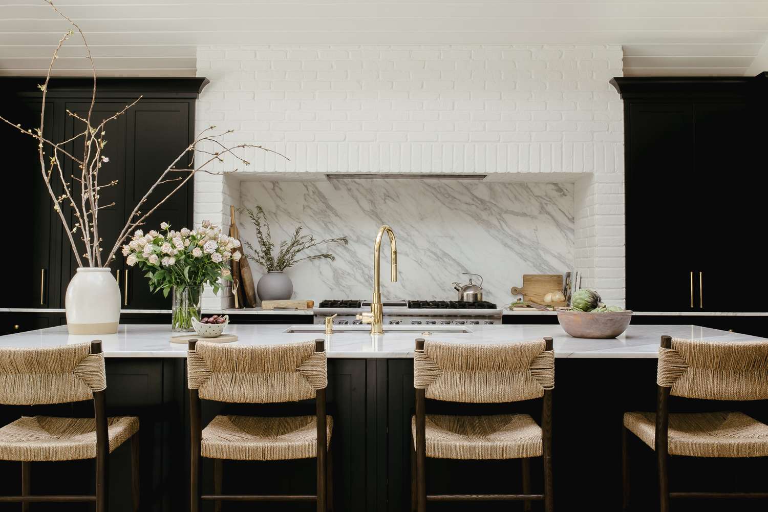 Modern kitchen interior with a marble backsplash, a countertop featuring a vase with tall branches and floral arrangements, and woven-seated chairs at a kitchen island