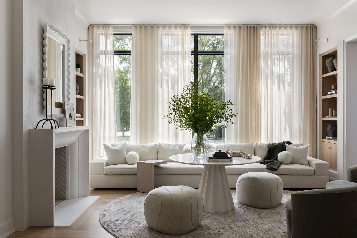 Minimalist living room by Brad Ramsey Interiors: creamy upholstered sofas with white pillows, marble fireplace, round glass coffee table, and potted greenery. Sheer curtains on large windows, light wood floor, and natural light create a calm, elegant space.