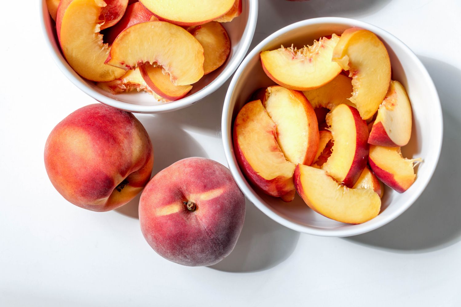 Whole ripe peaches on a white background next to bowls of sliced peaches. 