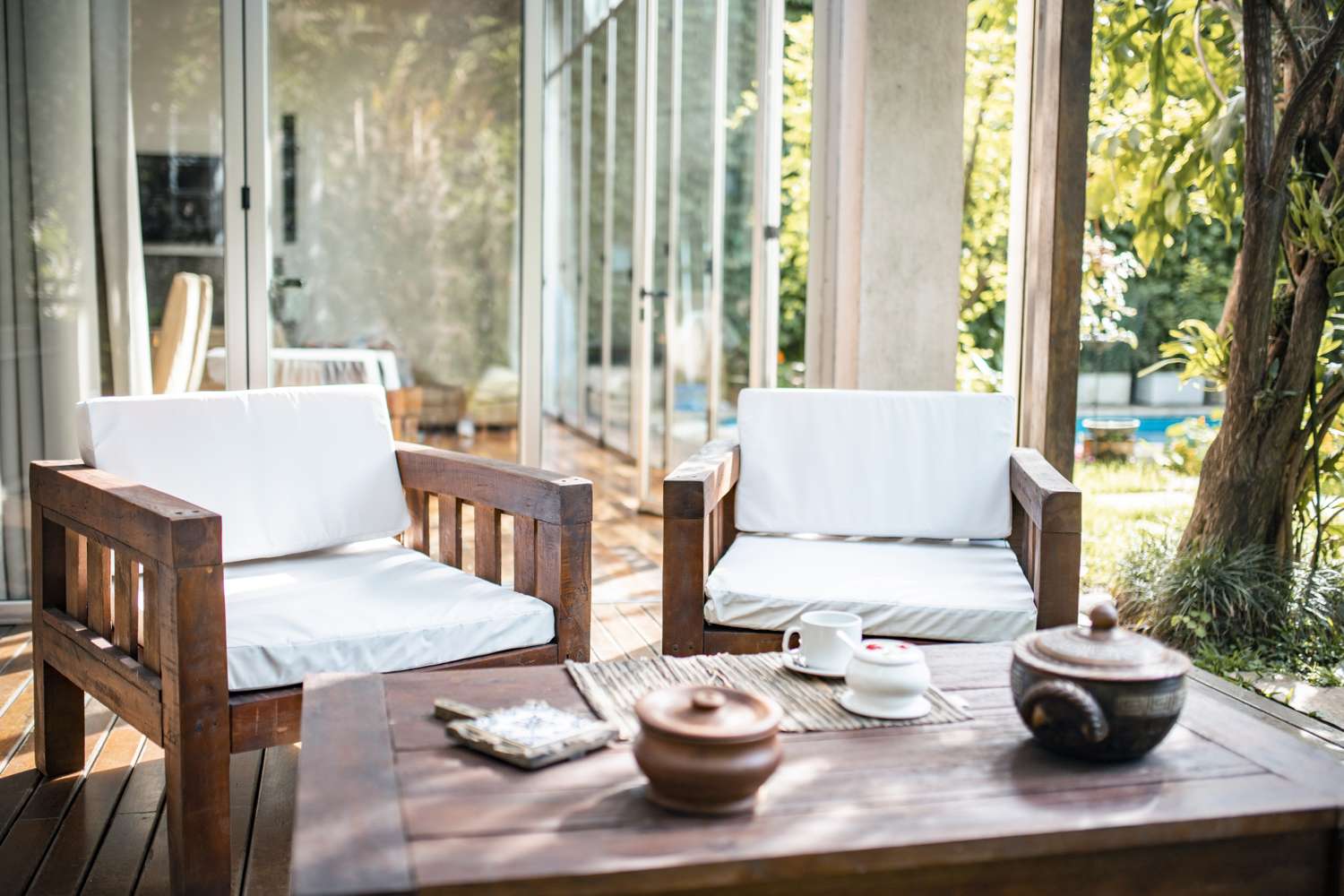 Outdoor patio with wooden furniture including two chairs and a table a tea set and pots on the table trees and greenery in the background