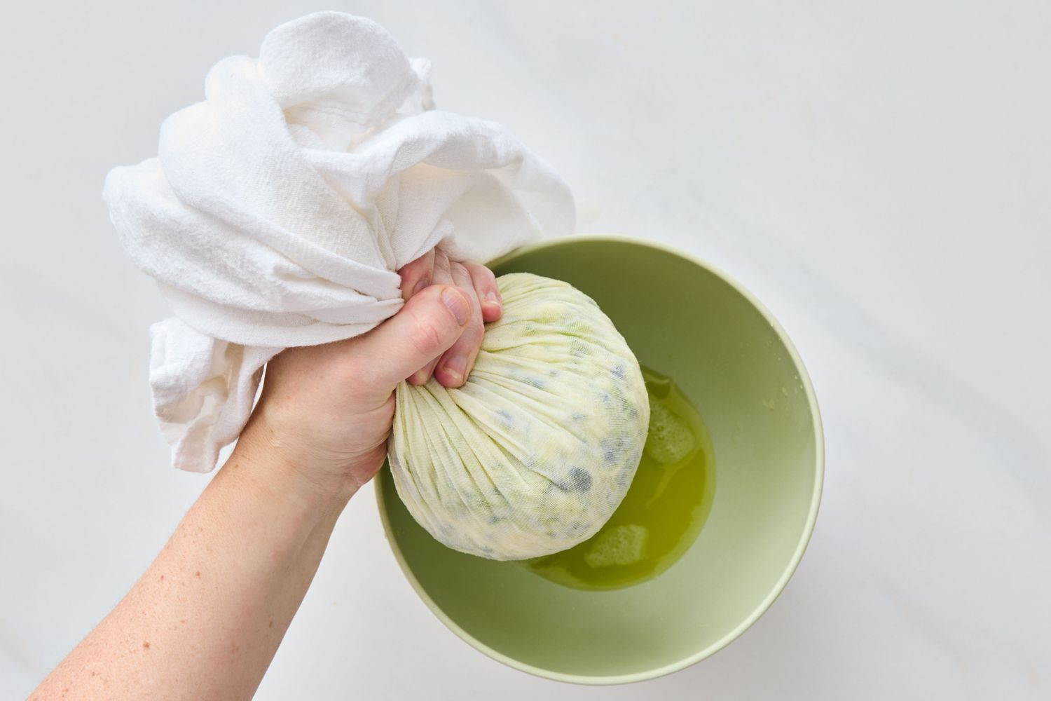A hand squeezing liquid from grated zucchini wrapped in a cloth into a green bowl