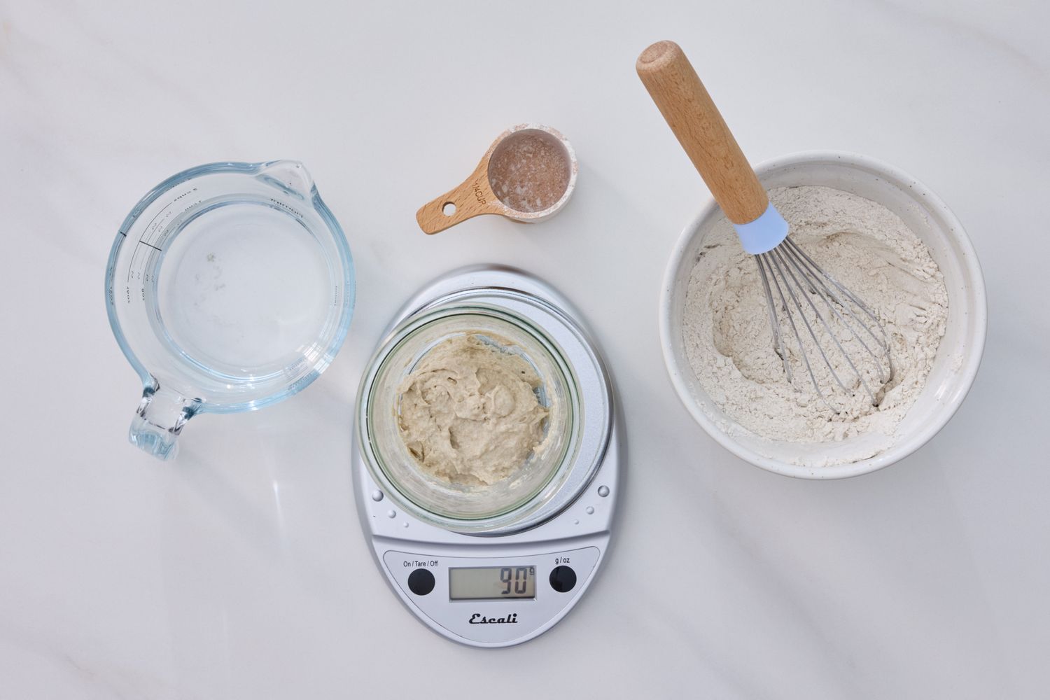 Ingredients and tools for making sourdough starter including flour water a scale and a whisk on a countertop