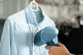 A hand using a steamer on a shirt hanging on a hanger steam visible near the shirt