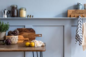 A kitchen counter scene with lemons artichokes in a basket a potted plant cutting boards and jars with a bluepaneled wall background