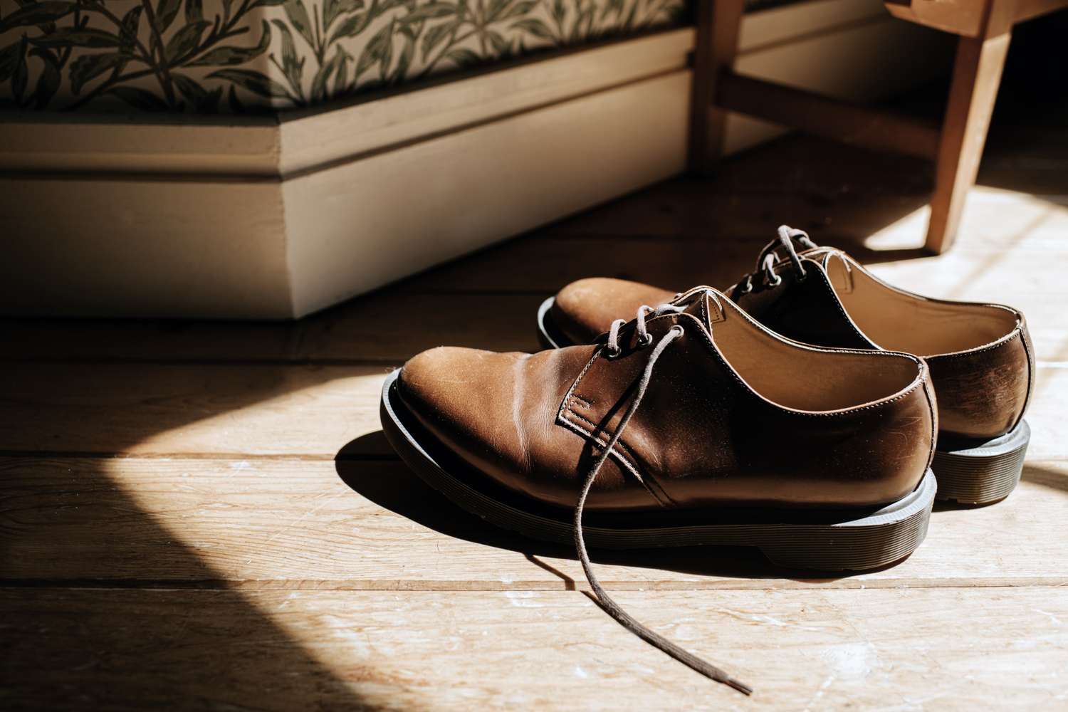 A pair of leather dress shoes on a wooden floor near a corner of a room with floral wallpaper