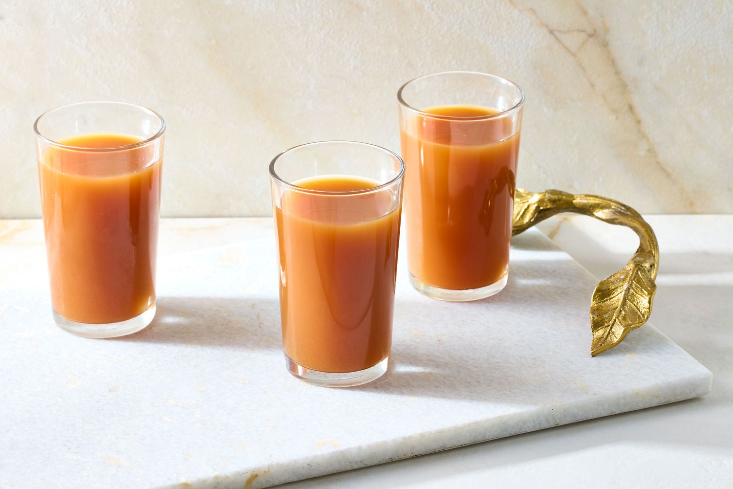 Three glasses of apple cider displayed on a marble surface accompanied by a decorative golden leaf