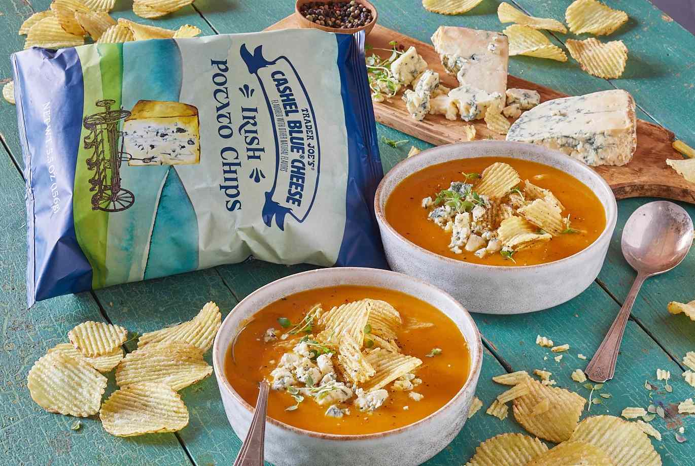 Two bowls of soup garnished with potato chips and cheese accompanied by a bag of Cashel Blue Cheese Irish Potato Chips and a cutting board with pieces of cheese on a table