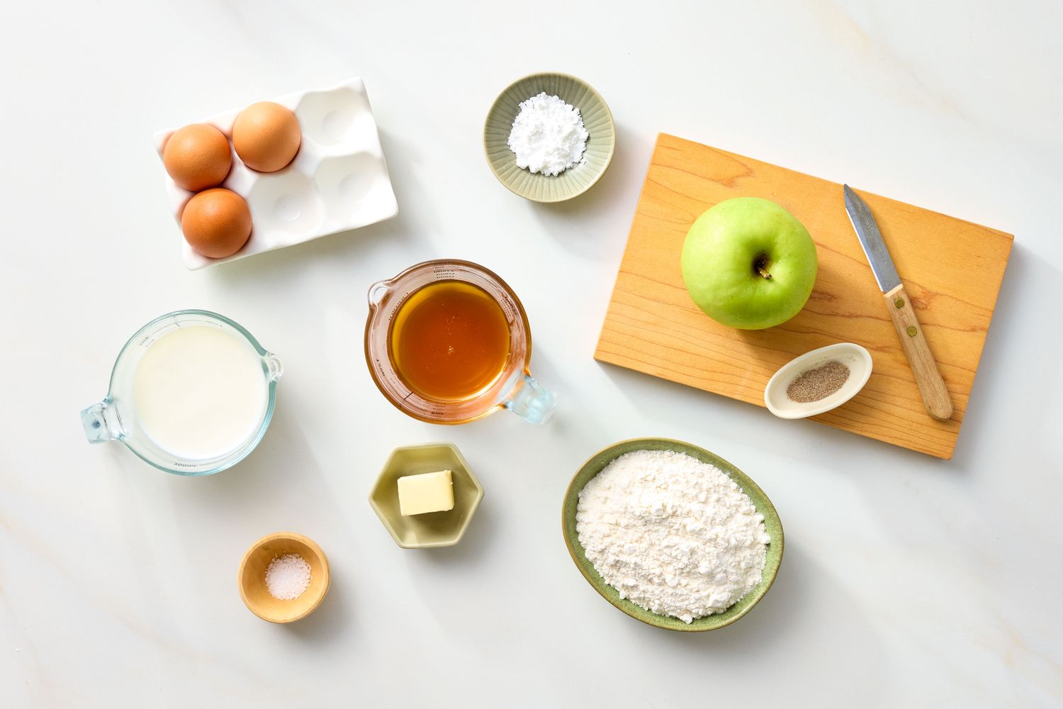 Ingredients for making a Dutch Baby pancake displayed on a countertop including eggs milk flour butter apple and seasonings