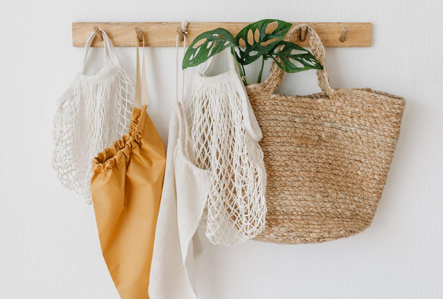 Reusable bags and a straw bag hanging on a wallmounted wooden rack with a few leaves above