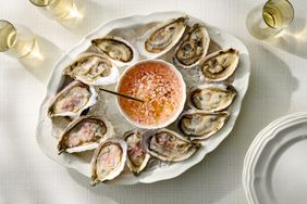 A platter of oysters on ice with a bowl of mignonette sauce in the center surrounded by glasses of a drink on a white tablecloth