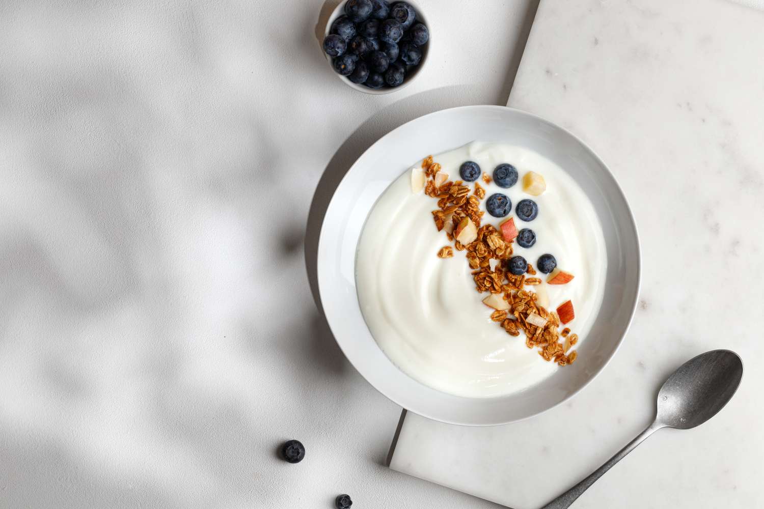 A bowl of yogurt topped with granola and blueberries a spoon and a small bowl of blueberries beside it