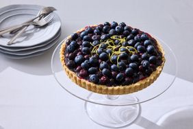 A blueberry tart garnished with lemon zest on a glass cake stand with a stack of plates and forks nearby