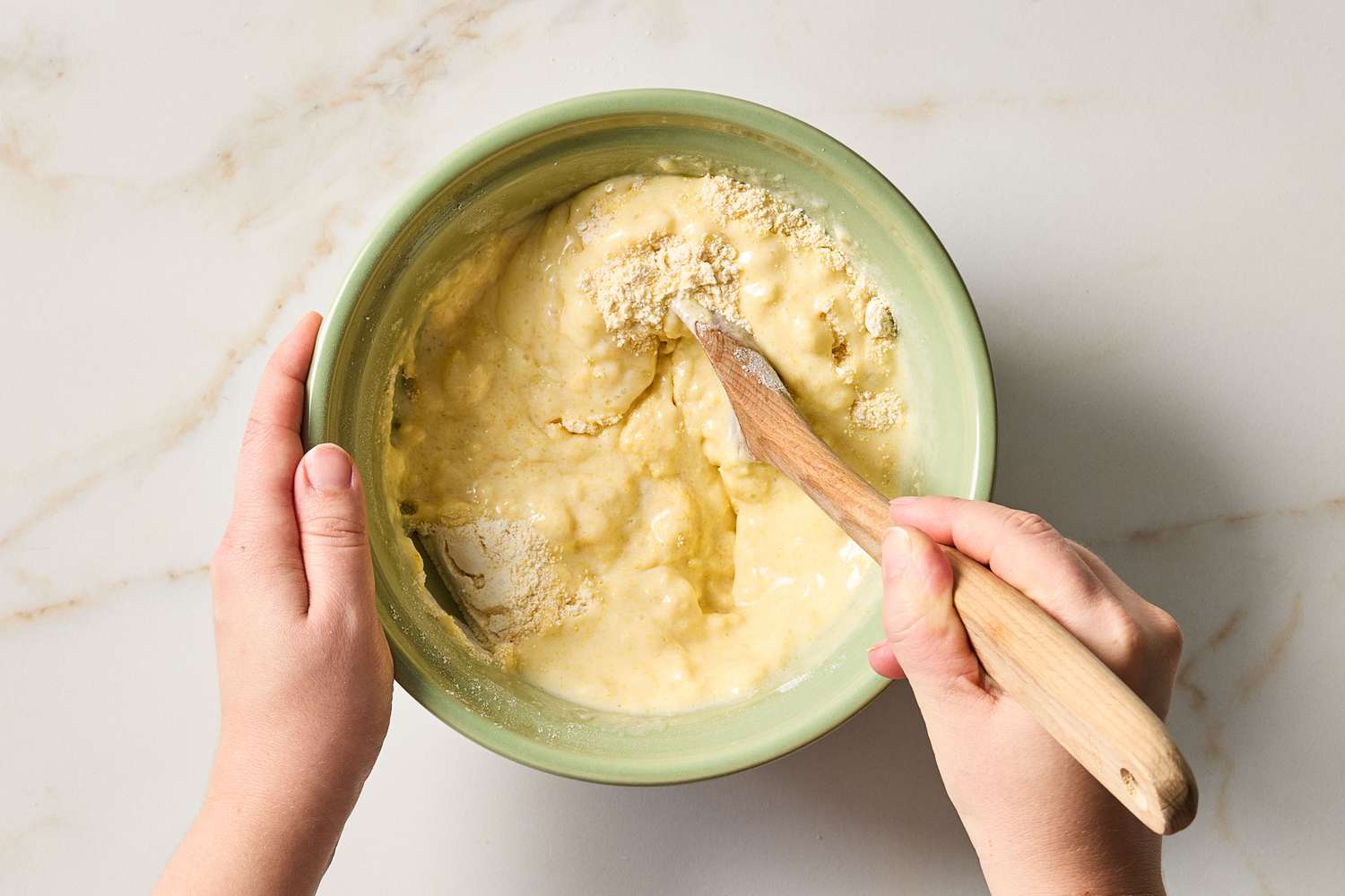 Hands stirring batter in a green bowl with a wooden spoon