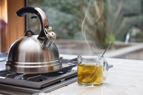 A stovetop kettle on a burner next to a steaming cup of tea on a tiled surface