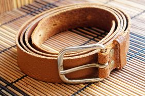 A rolled brown leather belt with a metal buckle on a bamboo mat