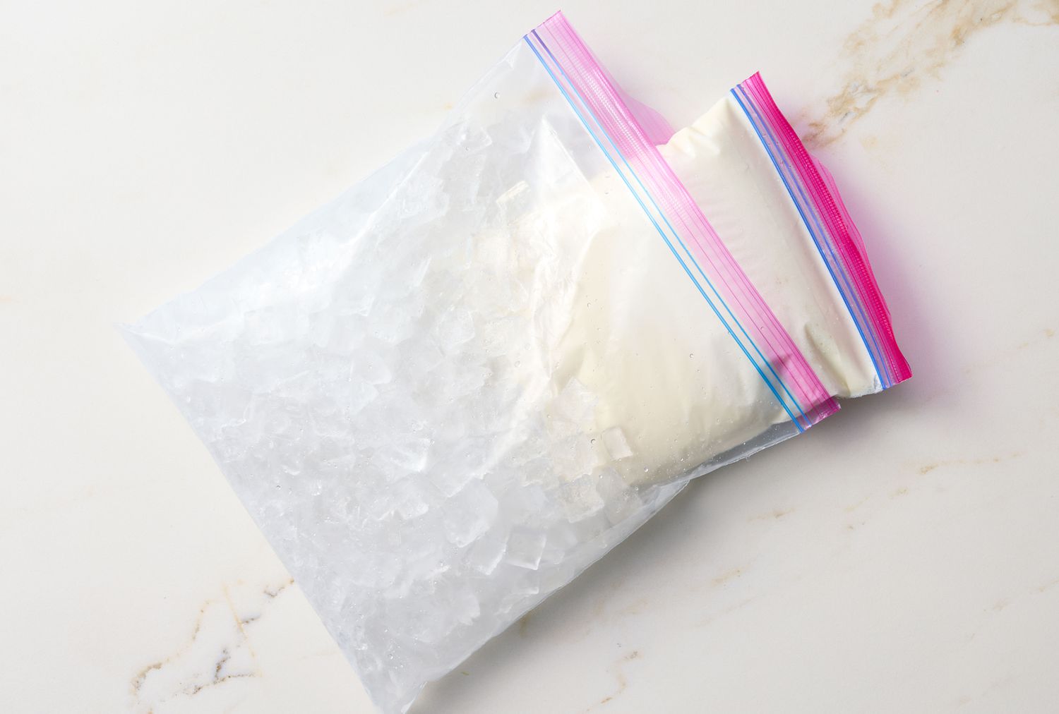 A plastic bag filled with ice and a smaller bag inside with a creamy mixture, placed on a light surface