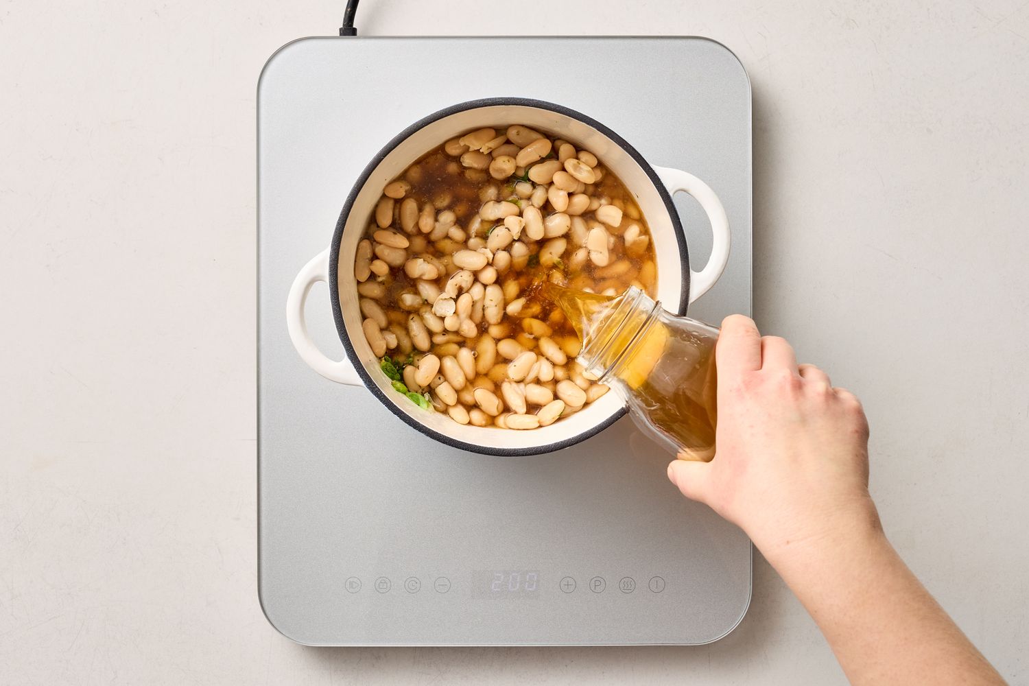 A hand pouring liquid into a pot of white beans on a stovetop