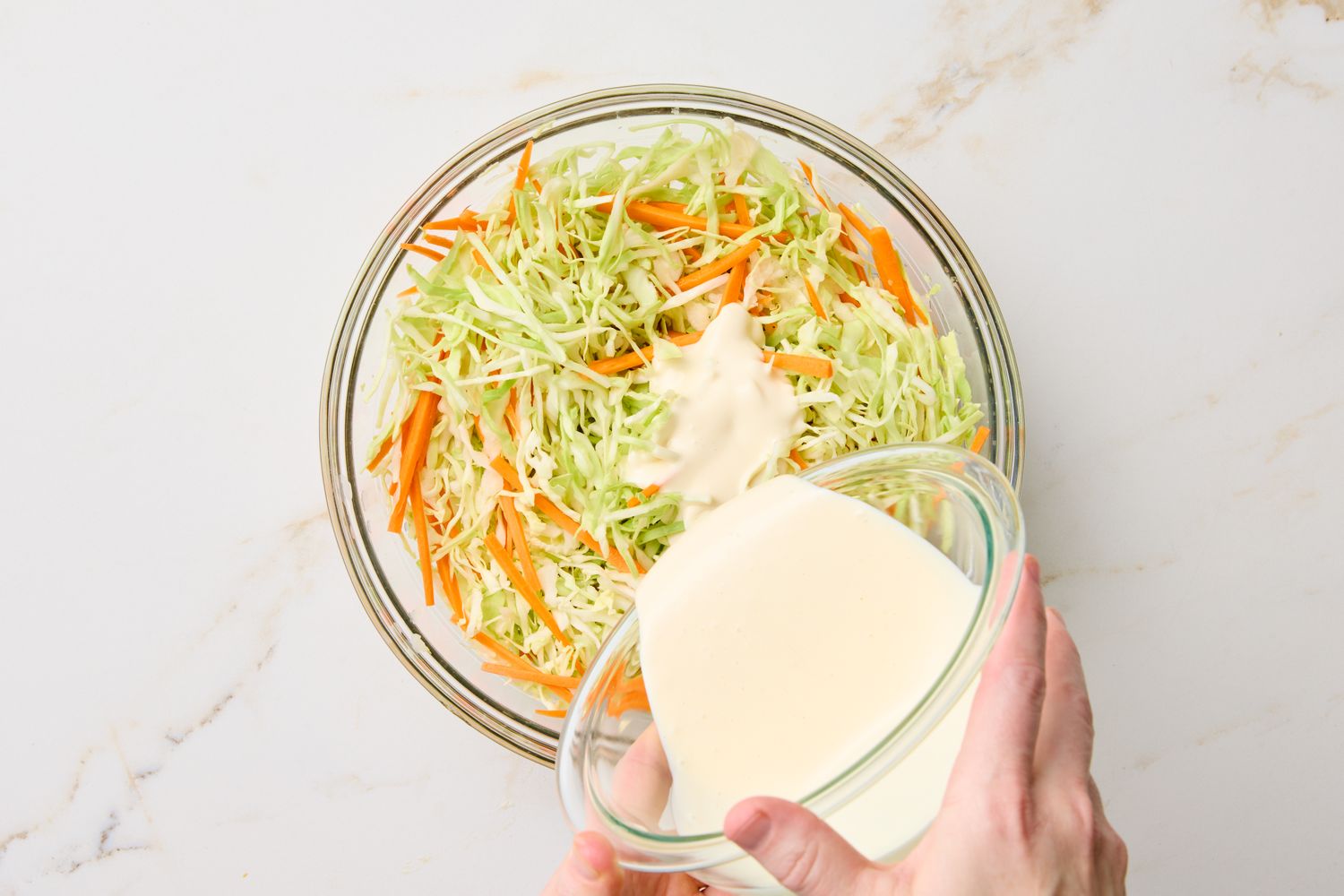 Hand pouring dressing onto a bowl of shredded cabbage and carrots for coleslaw