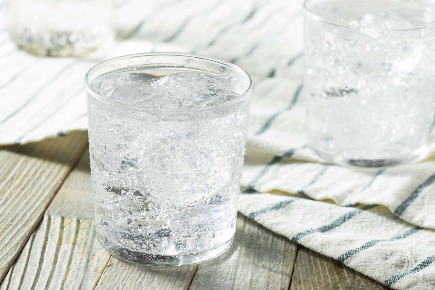 A glass of carbonated water with ice cubes placed on a wooden surface with a striped cloth
